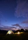 Burning bonfire near the tent in which light is on against backdrop of powerful mountains under starry sky