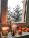 Burning candles in golden vintage candlesticks with group of mandarines at the window sill. Cozy winter day at home.