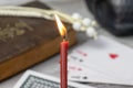 Burning church red candle in focus, blurred old holy bible, black skull and cards on wooden table. Misticism and fortune telling,
