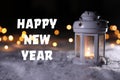 Burning lantern on snow and message HAPPY NEW YEAR against blurred background.