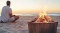 Burning logs in a rustic fire pit on a sandy beach. Person sits blurred in the distance by the ocean at sunset Royalty Free Stock Photo