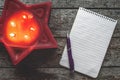 A burning red candle in the shape of a star and a blank paper with a pen lie on an old wooden table. Christmas wish list concept Royalty Free Stock Photo