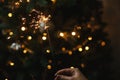 Burning sparkler in female hand on background of christmas tree lights in dark room. Happy New Year! Hand holding firework against Royalty Free Stock Photo