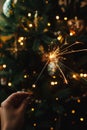 Burning sparkler in female hand on background of christmas tree lights in dark room. Happy New Year! Hand holding firework against Royalty Free Stock Photo