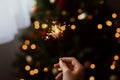 Burning sparkler in hand on background of golden bokeh lights in festive dark room. Hand holding firework at christmas tree with Royalty Free Stock Photo