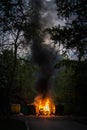 Burning trash container and old wood chair with other garbage at nght. Plastic garbage can has almost fused