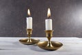 Burning two candles in vintage metal candlesticks on white wooden table against dark stone background. Royalty Free Stock Photo