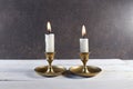 Burning two candles in vintage metal candlesticks on white wooden table against dark stone background. Royalty Free Stock Photo