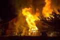 Burning wooden house at night. Bright orange flames and dense smoke from under the tiled roof on dark sky, trees silhouettes and r