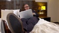 Business bald man with glasses and a stylish business suit, lying on the bed in the hotel room and looking at the laptop