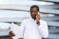 Business Call. Serious Black Businessman Talking On Cellphone And Reading Documents Outdoors Royalty Free Stock Photo