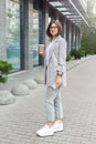 Business casual. Young lady in eyeglasses standing on the city street drinking coffee looking camera joyful full body