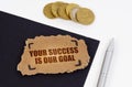 On a black and white surface lies a pen, coins and a cardboard sign with the inscription - Your Success is Our Goal