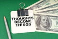 On a green surface, a pen, dollars and paper with the inscription - Thoughts Become Things