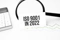 Business concept. Top view of calculator, magnifier, pen, table clock and notebook written ISO 9001 in 2022 on wooden background