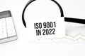 Business concept. Top view of calculator, magnifier, pen, table clock and notebook written ISO 9001 in 2022 on wooden background