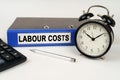 On a white surface, an alarm clock calculator and a folder with the inscription - LABOUR COSTS