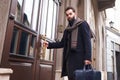 Business elegant man opens the door to his office. Stylish man in a coat with a beard enters the building with a bag in his hands. Royalty Free Stock Photo