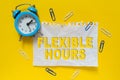 On the table lies an alarm clock, paper clips and a sheet of paper with the inscription - FLEXIBLE HOURS. Yellow background