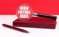 On a white and red background, a notebook, a pen and a magnifying glass, inside which the inscription - High Paying Jobs Royalty Free Stock Photo