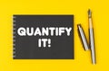 On a yellow background lies a pen and a black notebook with the inscription - Quantify It