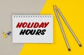 On a yellow-gray background are pencils, buttons and a notebook in which it is written - HOLIDAY HOURS