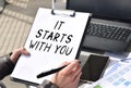 Business girl works at the table outdoors. The girl is holding a tablet for papers with the inscription IT STARTS WITH YOU.