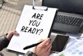 Business girl works at the table outdoors. The girl is holding a tablet for papers with the inscription ARE YOU READY. Conceptual