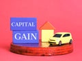 Text capital gain on wooden blocks with house and car miniature against red background. Royalty Free Stock Photo