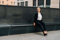 A business lady in a black, stern suit sits on a bench outside work at lunchtime. Customer`s waiting concept