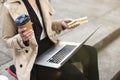 Business lady holding laptop on her knees with coffee and sandwich in hands working during lunch break on the stairs near office, Royalty Free Stock Photo