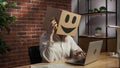 Portrait of female in cardboard box with emoji on head. Worker sitting at the desk working on laptop, talking on