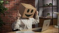 Portrait of female in cardboard box with emoji on head. Worker sitting at the desk looking at laptop, happy dancing