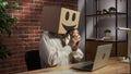 Portrait of female in cardboard box with emoji on head. Worker sitting at the desk working on laptop, holding cup of