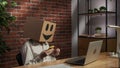 Portrait of female in cardboard box with emoji on head. Worker at the desk holding smartphone and credit card typing