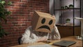 Portrait of female in cardboard box with emoji on head. Worker sitting at the desk looking at smartphone sad hopeless