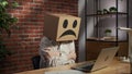Portrait of female in cardboard box with emoji on head. Worker sitting at the desk working on laptop, sad face feeling