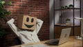 Portrait of female in cardboard box with emoji on head. Worker sitting at the desk working on laptop, leaning back on Royalty Free Stock Photo