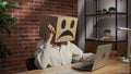 Portrait of a woman in a cardboard box with a negative emoji on her head. Employee sitting at desk, working on laptop Royalty Free Stock Photo