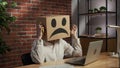 Portrait of a woman in a cardboard box with a negative emoji on her head. Employee sitting at desk, working on laptop Royalty Free Stock Photo