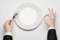 Business lunch and healthy food theme: man's hand in a black suit holding a white empty plate and shows finger gesture on an