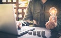 Business man hand holding lightbulb on stack coins, and working with computer on workplace. Creative ideas concept of saving money Royalty Free Stock Photo