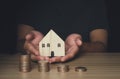 A business man holds a wooden house model in his hand and has a stack of coins money in front Royalty Free Stock Photo