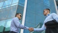 Business man passing a black briefcase to his partner. Colleagues shake hands outdoor in the city background. Two young Royalty Free Stock Photo