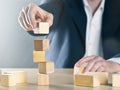 Business man puts next stone on shaky tower structure made from wooden blocks; career or achievement or complex project management