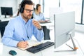 Business man wearing headset looking at computer and taking notes to notebook accompanied with colleagues in background. Royalty Free Stock Photo