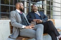 Business meeting of friends outdoors. Two dark-skinned men in suits are sitting on a bench near a city building with a Royalty Free Stock Photo