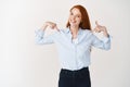 Business people. Skillful young redhead woman pointing at herself and smiling, being a professional, standing over white Royalty Free Stock Photo