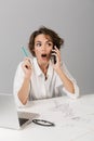 Business shocked woman posing isolated over grey wall background sitting at the table using laptop drawing talking by phone