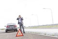 Businessman talking on phone while standing with breakdown car by warning sign on road Royalty Free Stock Photo